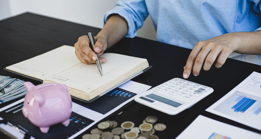 Imagen de una persona escribiendo en un cuaderno y usando una calculadora junto a una alcancía en forma de chanchito para gestionar sus finanzas, usada en una entrada del blog de Chapa Cambio sobre métodos prácticos para ahorrar dinero