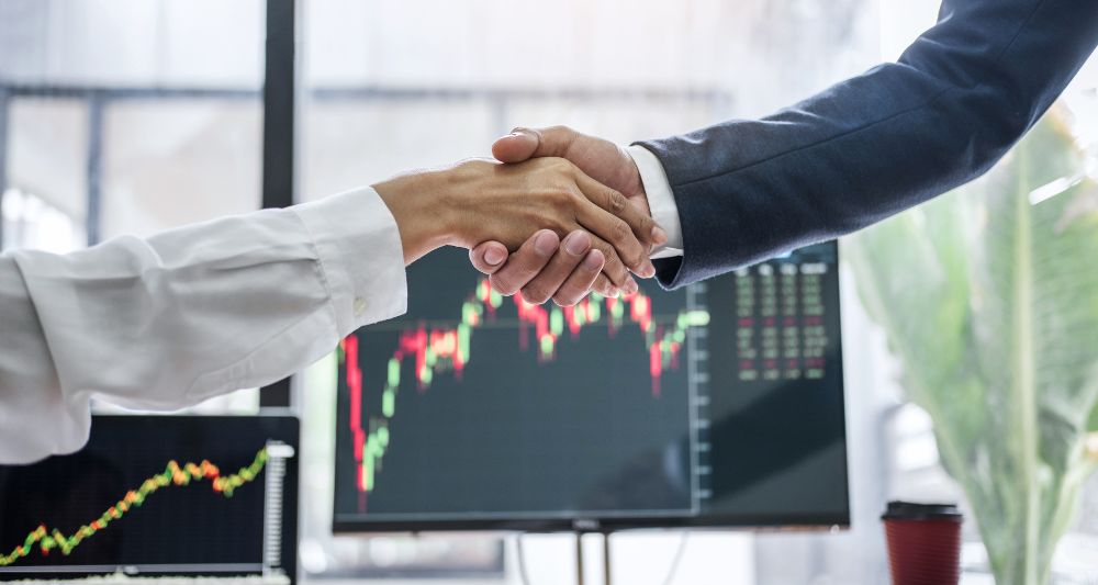 Imagen de dos personas dándose la mano frente a pantallas con gráficos financieros, usada en una entrada en el blog de Chapa Cambio sobre cuál es cómo elegir al mejor broker del Perú según tus necesidades