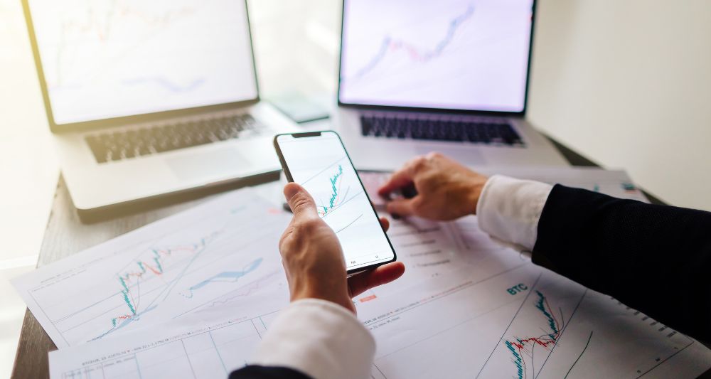 Foto mostrada en una entrada del blog de Chapa Cambio sobre cómo comprar acciones en el Perú que presenta a una persona revisando gráficos y datos financieros en varios dispositivos en medio de una investigación y análisis necesarios para la compra de acciones