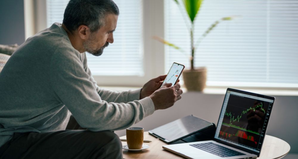 Foto mostrada en una entrada del blog de Chapa Cambio sobre cómo escoger acciones para invertir que muestra a un hombre analizando gráficos de acciones en su laptop y smartphone mostrando el proceso de investigación y selección de acciones para realizar inversiones inteligentes